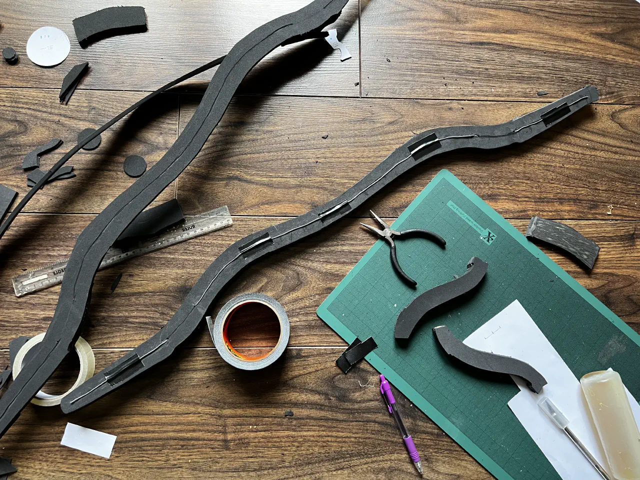 The handle of the bow lying on the floor with wire taped to it matching the shape. At two points the wire goes into the foam and comes out about 5cm along. Above it is the other side of the bow handle showing a shallow groove cut in to hold the wire.