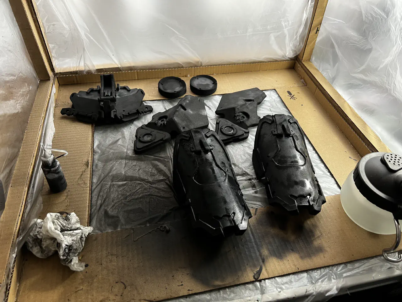 Seven primed pieces of armour sit in a cardboard framed box, the walls covered with plastic sheeting and splattered with black paint. 