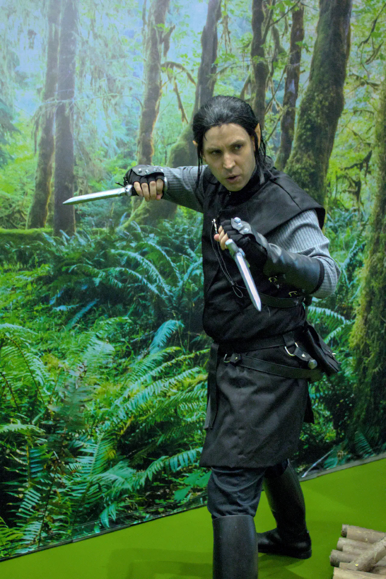 A individual in a black and grey outfit with black hair and pointed ears looking to one side whilst holding two knives