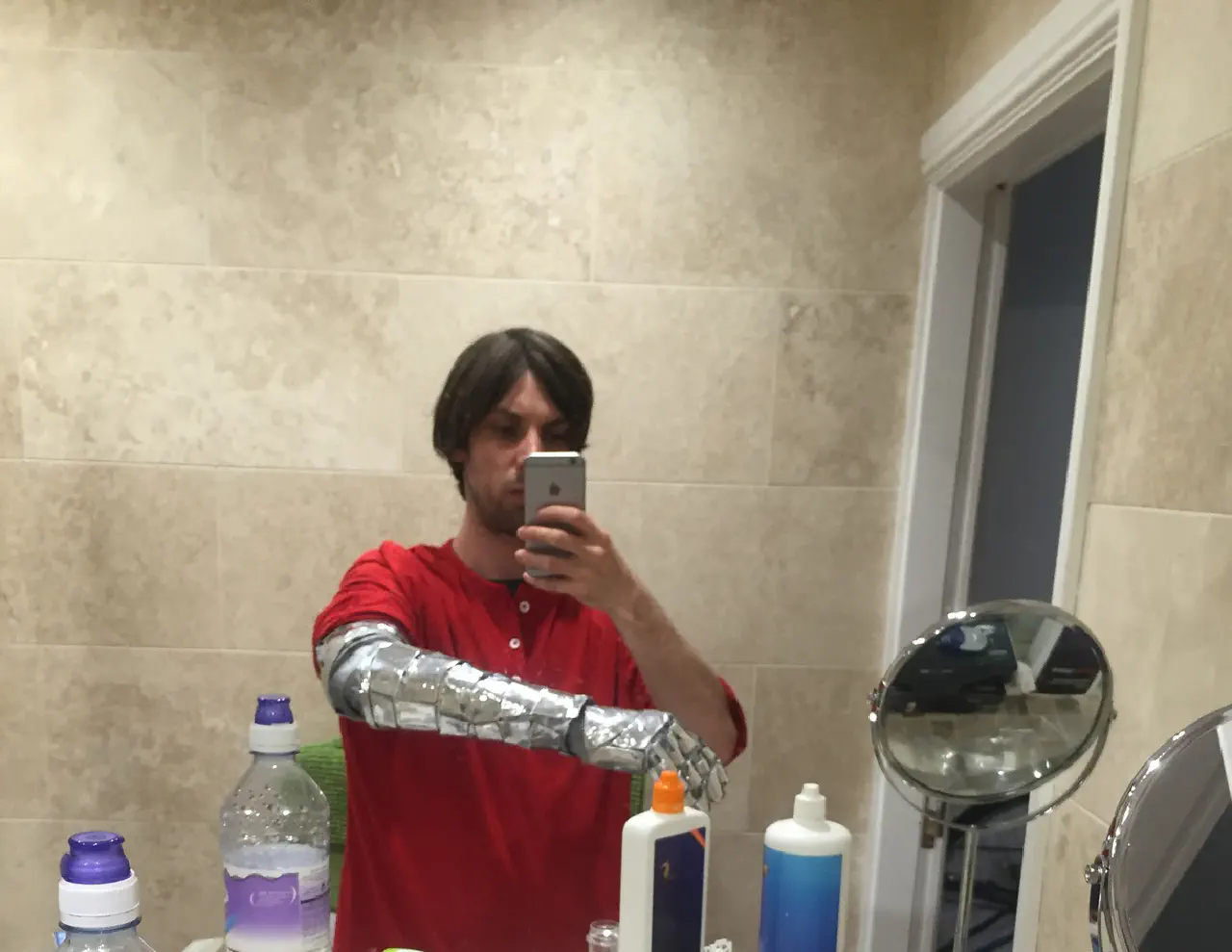 A stubbled individual in a read shirt taking a selfie in a large mirror. They have a shiny silver mechanical arm
