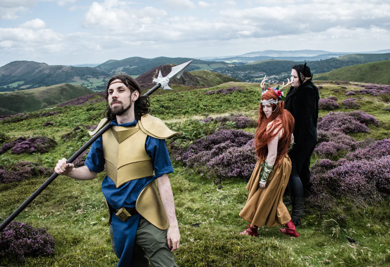 Three individuals standing on a grassy hill. The closest to the camera ia dressed in golden armour and holding a spear, the two further away are in fantasy costumes.