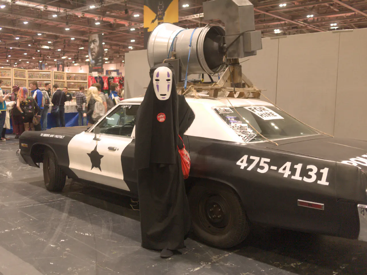 A figure draped in black obscuring any detail with a white and purple mask. They are standing in front of an old american style police car.