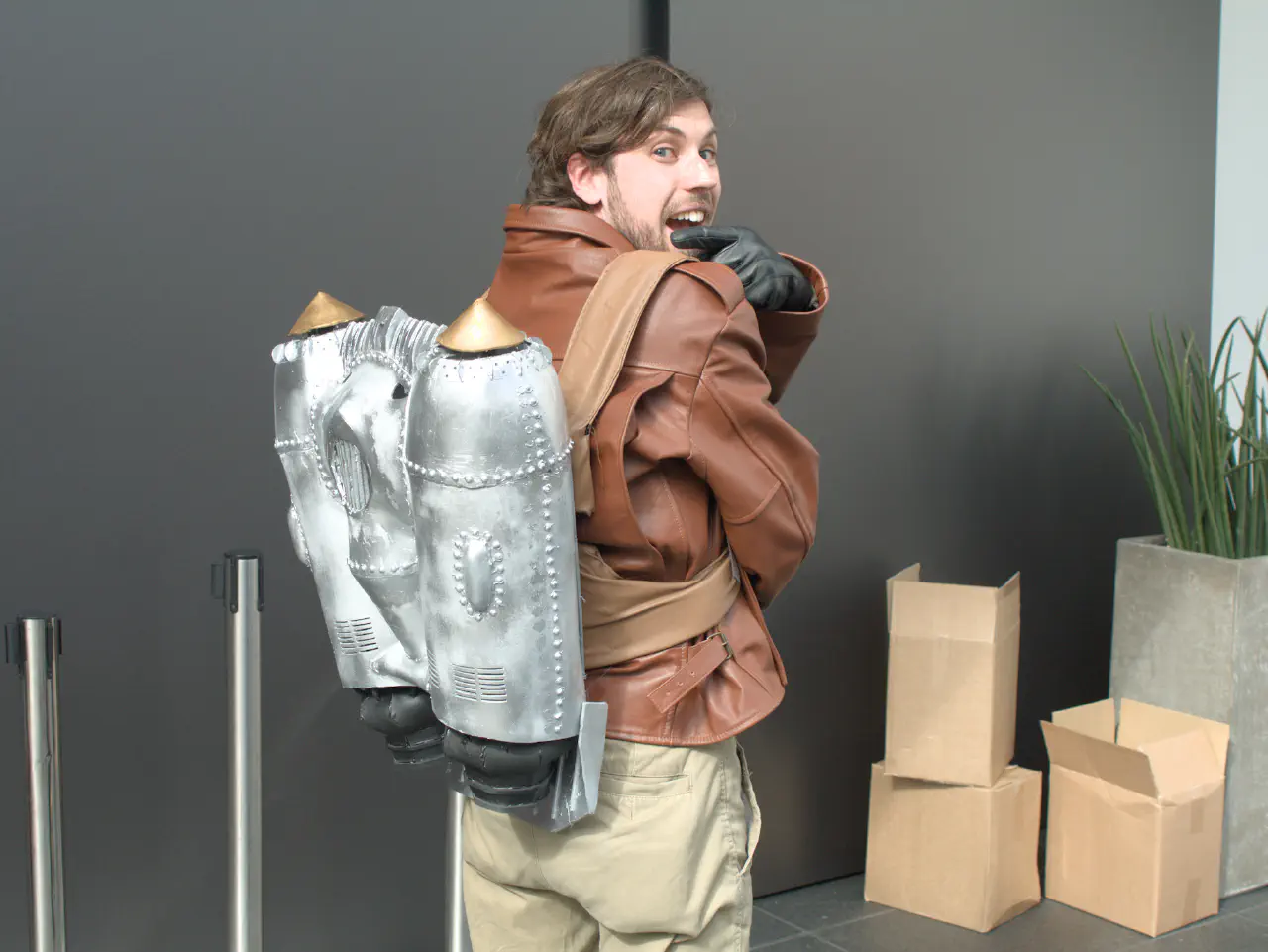 A bearded individual in a brown leather jacket facing away from the camera, but turning around to point very enthusiastically at the rocket pack they are wearing on their back.