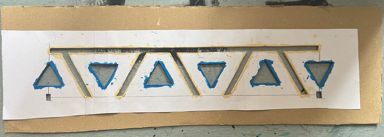 A line and vee template, cut out of paper and stuck to cardboard with a wide border. It is blotched with off white and blue paint.